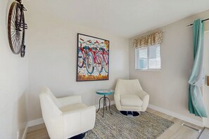 A room with two white armchairs, a small round table, a colorful bicycle painting, a window with blinds, and a curtain. - Bear Lake  View - Pool, Hot Tub & Game Room (Garden City)