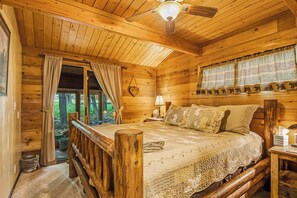 Une chambre à coucher dans une cabane en bois, avec un lit, une table de chevet, une lampe, une fenêtre avec des rideaux et un ventilateur de plafond.