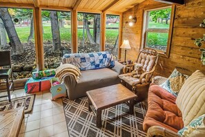 Ein gemütliches Zimmer mit einer Couch, einem Sessel und einem kleinen Tisch, großen Fenstern mit Blick auf Bäume und einer Holzbalkendecke.