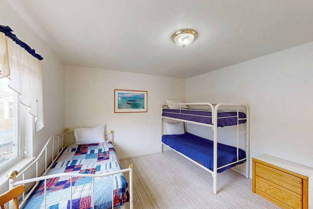 A room with a bunk bed, a single bed, a window with curtains, a framed picture on the wall, and a ceiling light fixture.