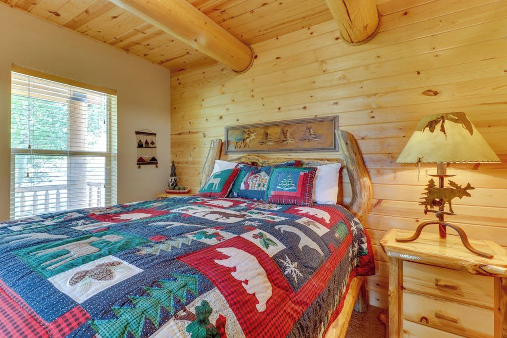 A wooden cabin bedroom with a large bed, a nightstand with a lamp, and a window with blinds.