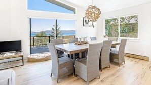 Ett modernt matområde med ett stort bord och stolar, en tv på ett stativ och utsikt över havet genom stora fönster.