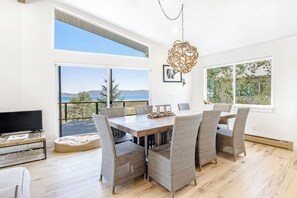 Un'area pranzo moderna con un tavolo grande e sedie, una televisione su un mobile e vista mare attraverso ampie finestre.