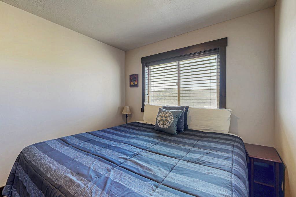 A bedroom with a bed, a window with blinds, a nightstand, and a wall-mounted lamp.