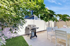 Eine Terrasse mit einem weißen Tisch und Stühlen, einem Grillwagen und einem weißen Zaun.