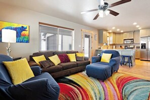A living room with a sofa, ottoman, and colorful rug. There is a ceiling fan, a window with blinds, and a kitchen in the background. - Cubbyz Cabin - Lake Views, Hot Tub, Near Marina (Garden City)