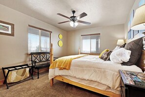 Ein Schlafzimmer mit einem Bett, einem Deckenventilator, zwei Fenstern mit Jalousien, einer Bank und einem Tisch mit einer Lampe.