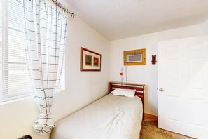 A single bed with a wooden headboard, a window with white curtains, a wall-mounted air conditioning unit, and a closed door. - Kokomo Kottage - Game Room, Fire Pit & Playset (Garden City)