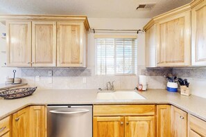 A kitchen with wooden cabinets, a stainless steel dishwasher, and a window with blinds. - Kokomo Kottage - Game Room, Fire Pit & Playset (Garden City)