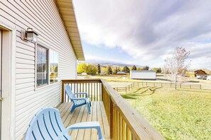 A wooden deck with two chairs, a view of a fenced yard, and a building with a window and a light fixture. - Kokomo Kottage - Game Room, Fire Pit & Playset (Garden City)