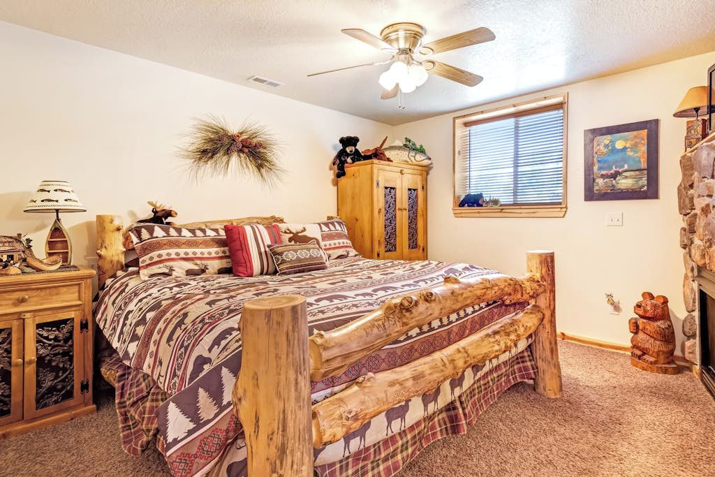 A bedroom with a wooden bed frame, a ceiling fan, a window with blinds, and a fireplace.