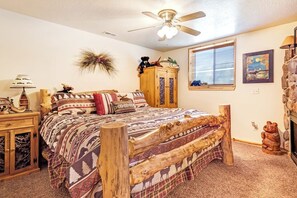 A bedroom with a wooden bed frame, a ceiling fan, a window with blinds, and a fireplace. - Moose Mountain Retreat (Fish Haven)