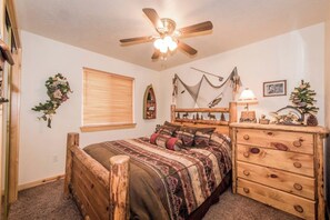A bedroom with a bed, a wooden dresser, a ceiling fan, a window with blinds, and a wall-mounted decoration. - Moose Mountain Retreat (Fish Haven)