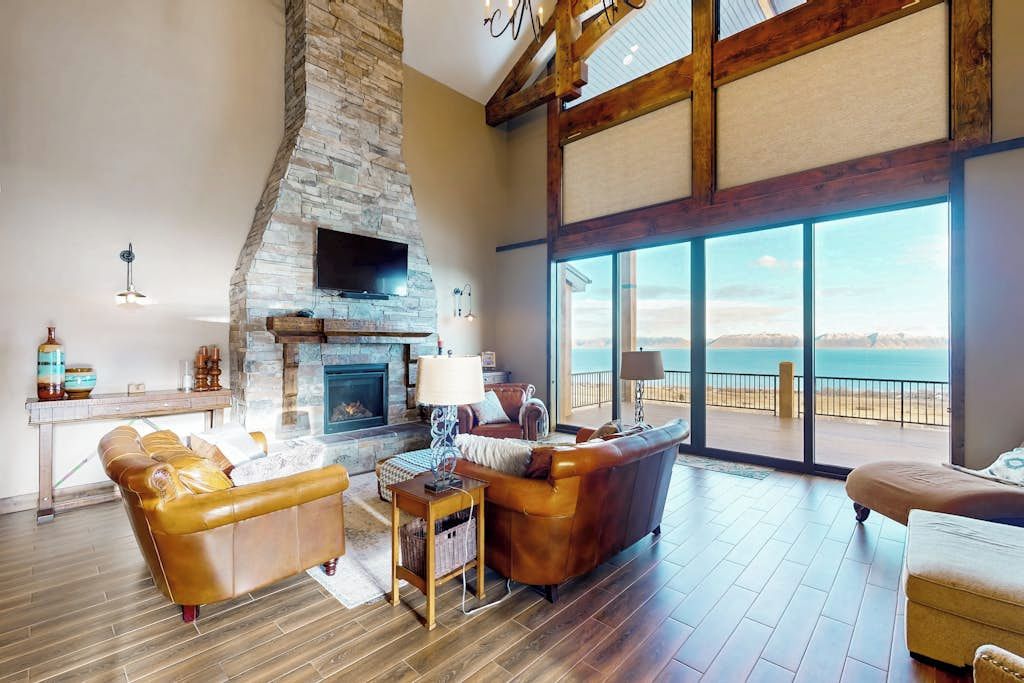 A spacious living room with a stone fireplace, leather sofas, and a large window overlooking a beach.