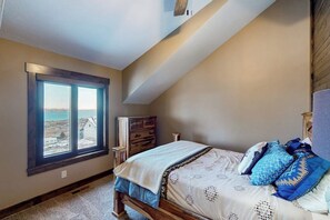 Une chambre avec une grande fenêtre, un lit recouvert d’une literie à motifs, une commode et une vue sur la mer.