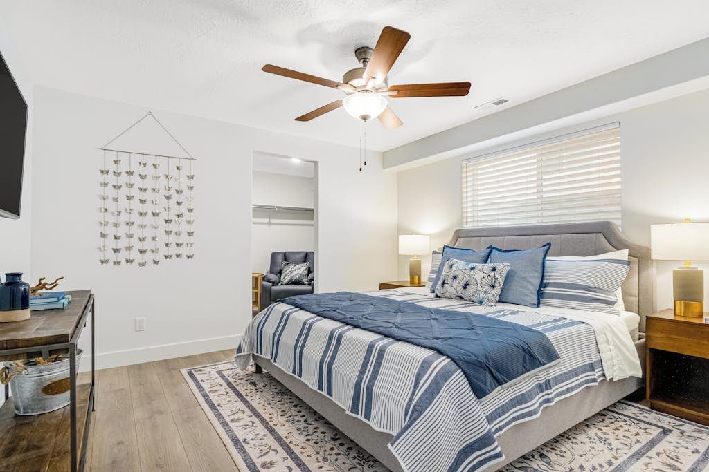 A bedroom with a bed, a ceiling fan, a wall-mounted light fixture, a window with blinds, and a decorative wall hanging.