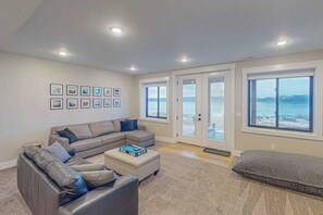 A living room with a sectional sofa, a small ottoman, and a grey couch. There are framed pictures on the wall and a large window with a view of the sea. - Bear Lake Retreat - Hot Tub & Views, Sleeps 18 (Garden City)