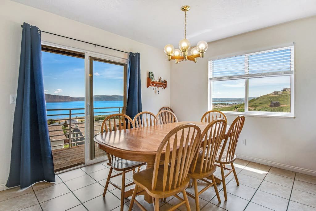 A dining area with a large table and chairs, a chandelier, a view of the sea through a sliding door, and a window with a view of the landscape.