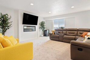 A modern living room with a flat-screen TV mounted on the wall, a brown leather sofa, a yellow armchair, and a large window with blinds. - Western Paradise at BearLake (Garden City)