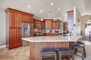 A kitchen with wooden cabinets, a stainless steel refrigerator, a microwave, and a granite countertop island with bar stools. - The Lakehouse - Lake Views, Sleeps 31, Dogs OK (Garden City)