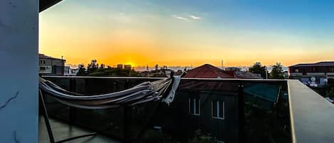 Un balcón con una hamaca, con vista a un paisaje urbano al atardecer.