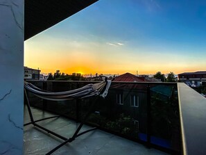 A balcony with a hammock, overlooking a cityscape during sunset.