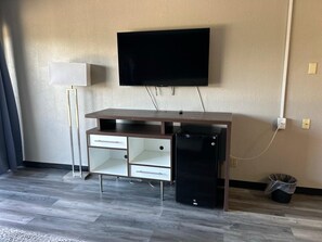 A room with a wooden desk, a flat-screen TV mounted on the wall, a small black refrigerator, and a trash bin.