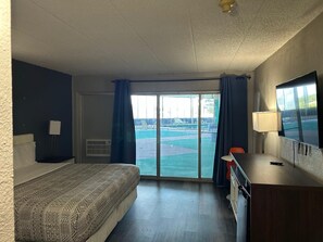 A hotel room with a bed, a desk, a TV, and a view of a pool and tennis court.