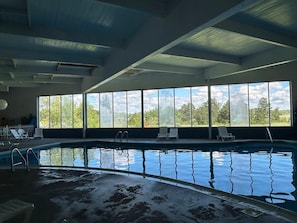 An indoor swimming pool with clear windows, lounge chairs, and a view of trees.