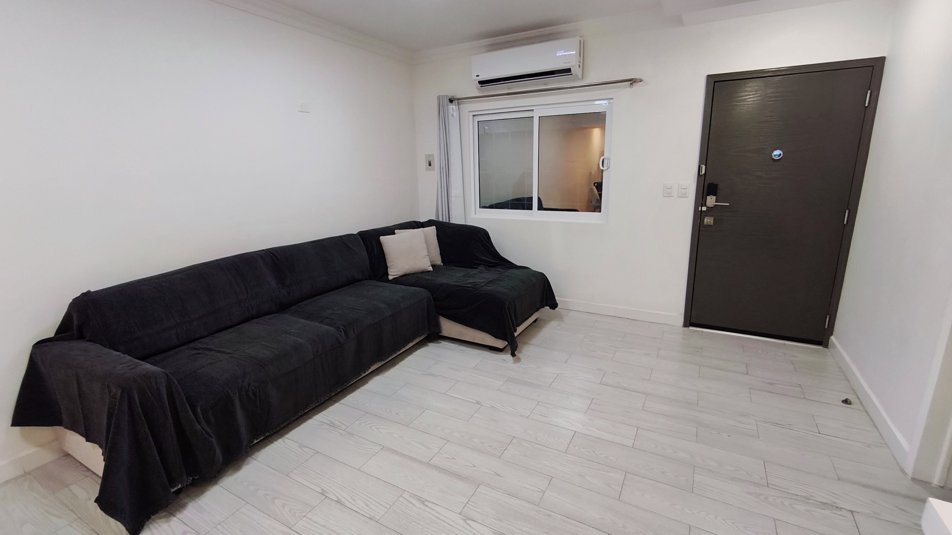 Una habitación con un sofá negro, una ventana con vistas, una puerta y un equipo de aire acondicionado.