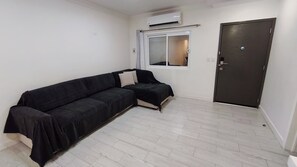 A room with a black sofa, a window with a view, a door, and an air conditioning unit.