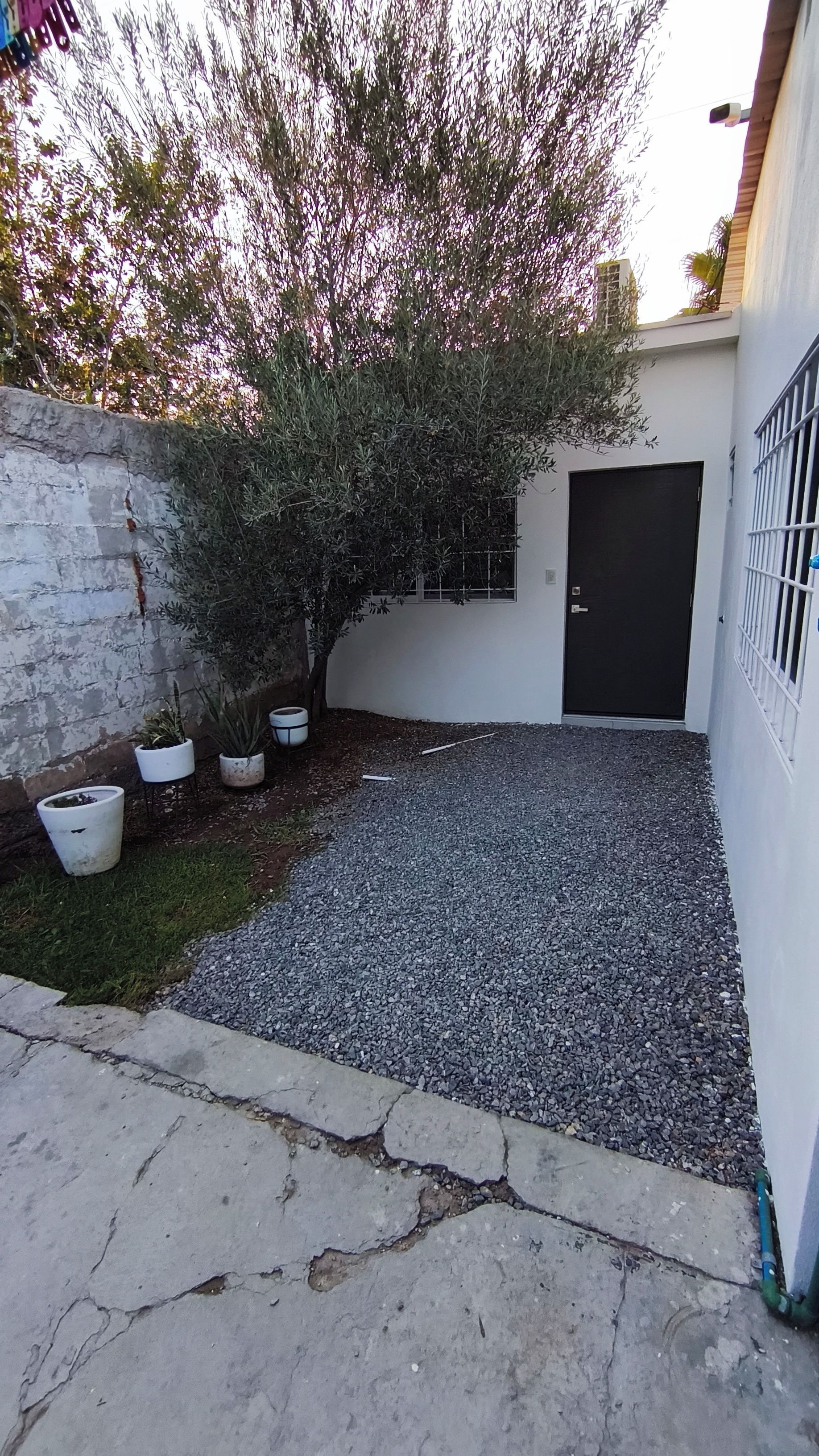 Un patio empedrado con un sendero de grava, un pequeño espacio con césped y una pared blanca con una puerta negra.