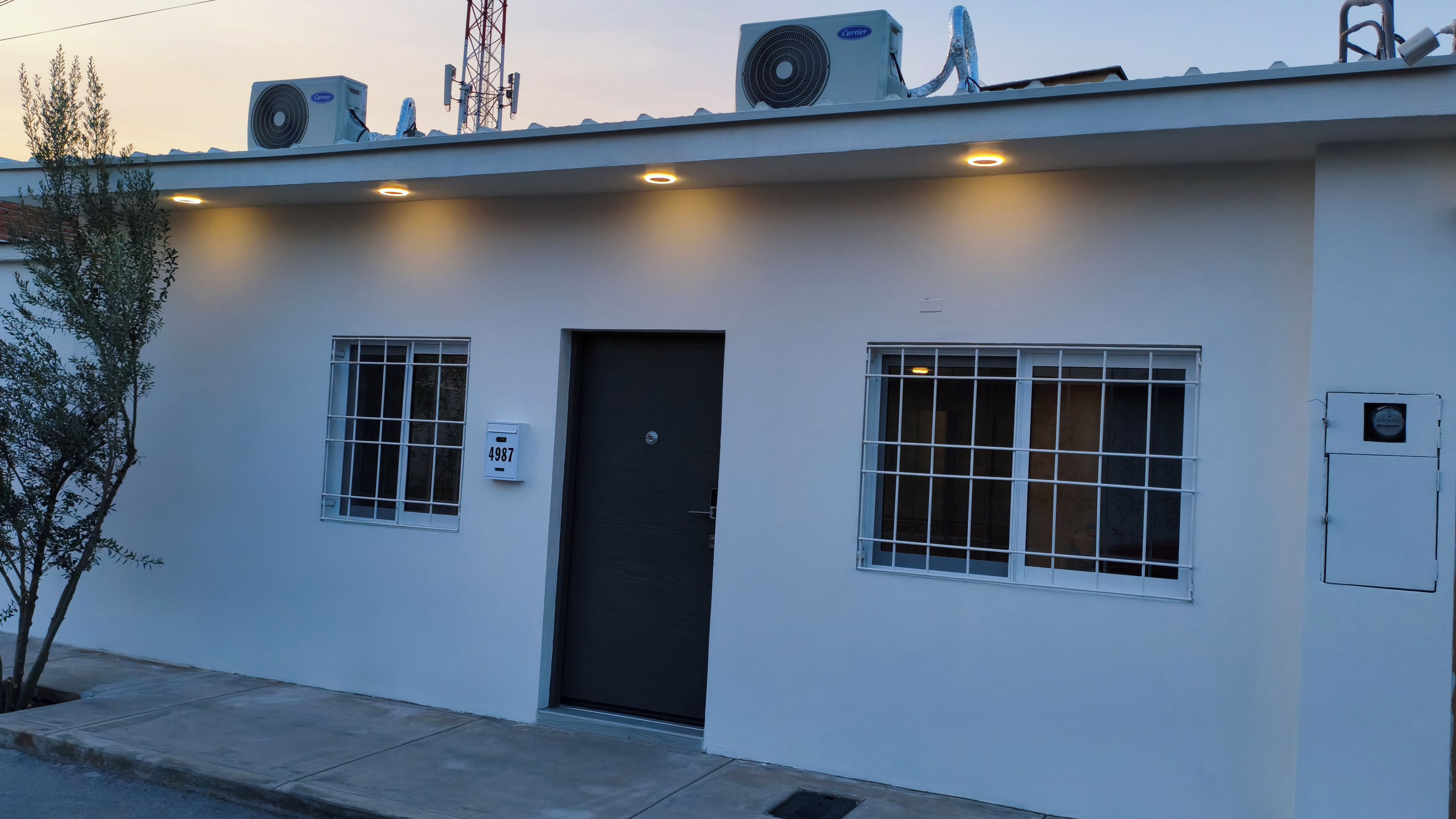 Un edificio con una puerta negra, dos ventanas con rejas y una placa con el número 4987 en la pared.