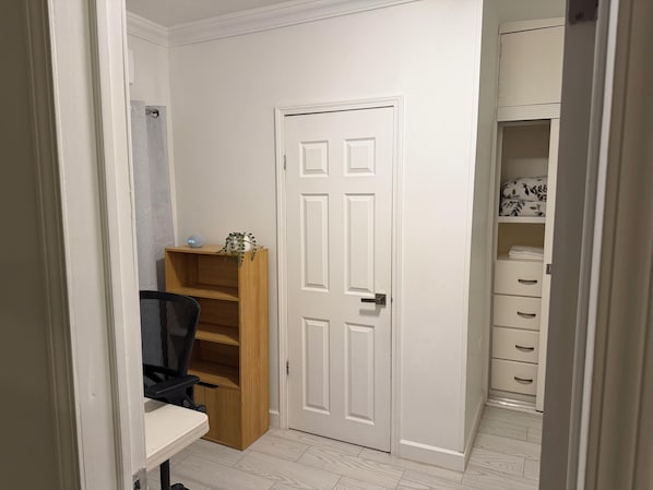 Un pasillo con una puerta blanca, una estantería de madera, una silla de oficina negra y un armario blanco con cajones.