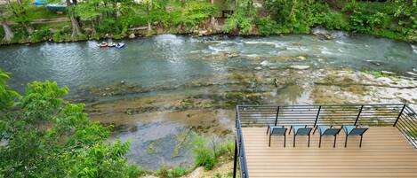Ein Fluss mit klarem Wasser, umgeben von üppigem Grün und einer Holzterrasse mit Stühlen.