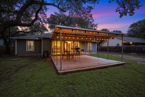Un'area patio coperta con mobili da esterno e luci a stringa, adiacente a una casa con garage e recinzione in legno.