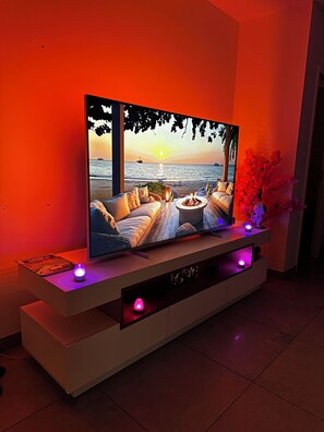 A flat-screen TV on a white entertainment unit with a sunset view on the screen, a small table with a lamp, and a vase with pink flowers. - Romantic getaway with a private hot tub, 15 minutes from Giverny (Freneuse)