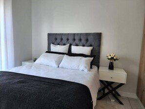 A neatly made bed with a dark headboard and white bedding, a bedside table with a vase of flowers, and a clean, minimalist room design. - Lovely One-bedroom Unit With Pool and Gym Facilities in Stand (Cape Town)
