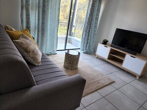 A modern living room with a sofa, a television on a wooden stand, and a view through large windows. - Lovely One-bedroom Unit With Pool and Gym Facilities in Stand (Cape Town)