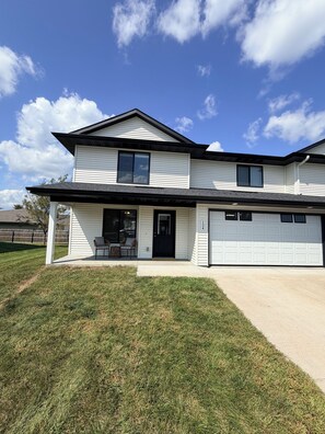 A two-story house with a garage, a front porch with seating, and a well-maintained lawn. - 3-bedroom townhome -8 Miles From Downtown Iowa City (Hills)