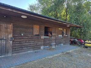Een houten stal met een paard zichtbaar door een geopende staldeur, een rood voertuig geparkeerd ervoor, en een verhard pad naar de ingang.