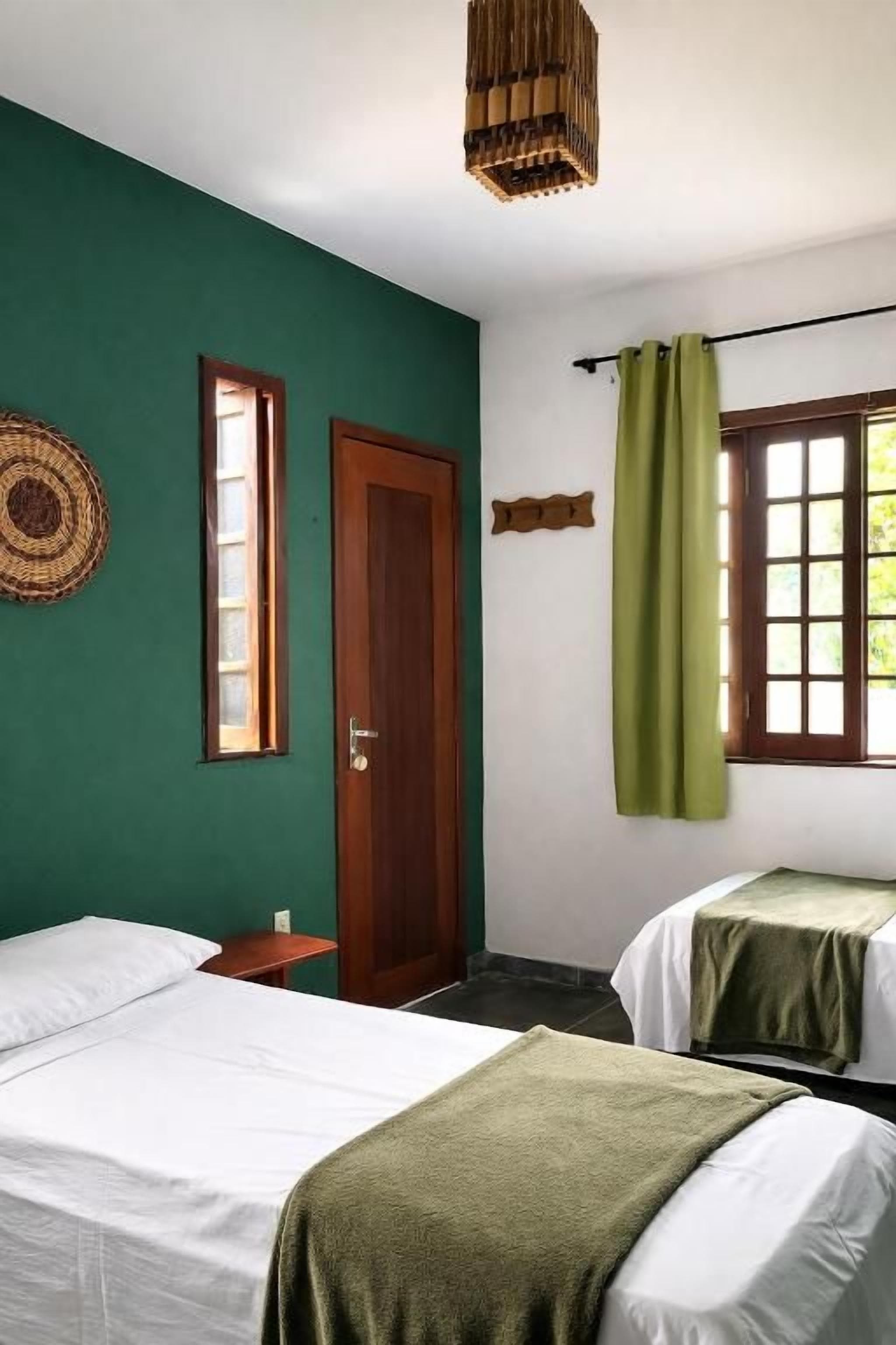 Een kamer met twee bedden, een houten deur, een raam met groene gordijnen en een geweven lamp.