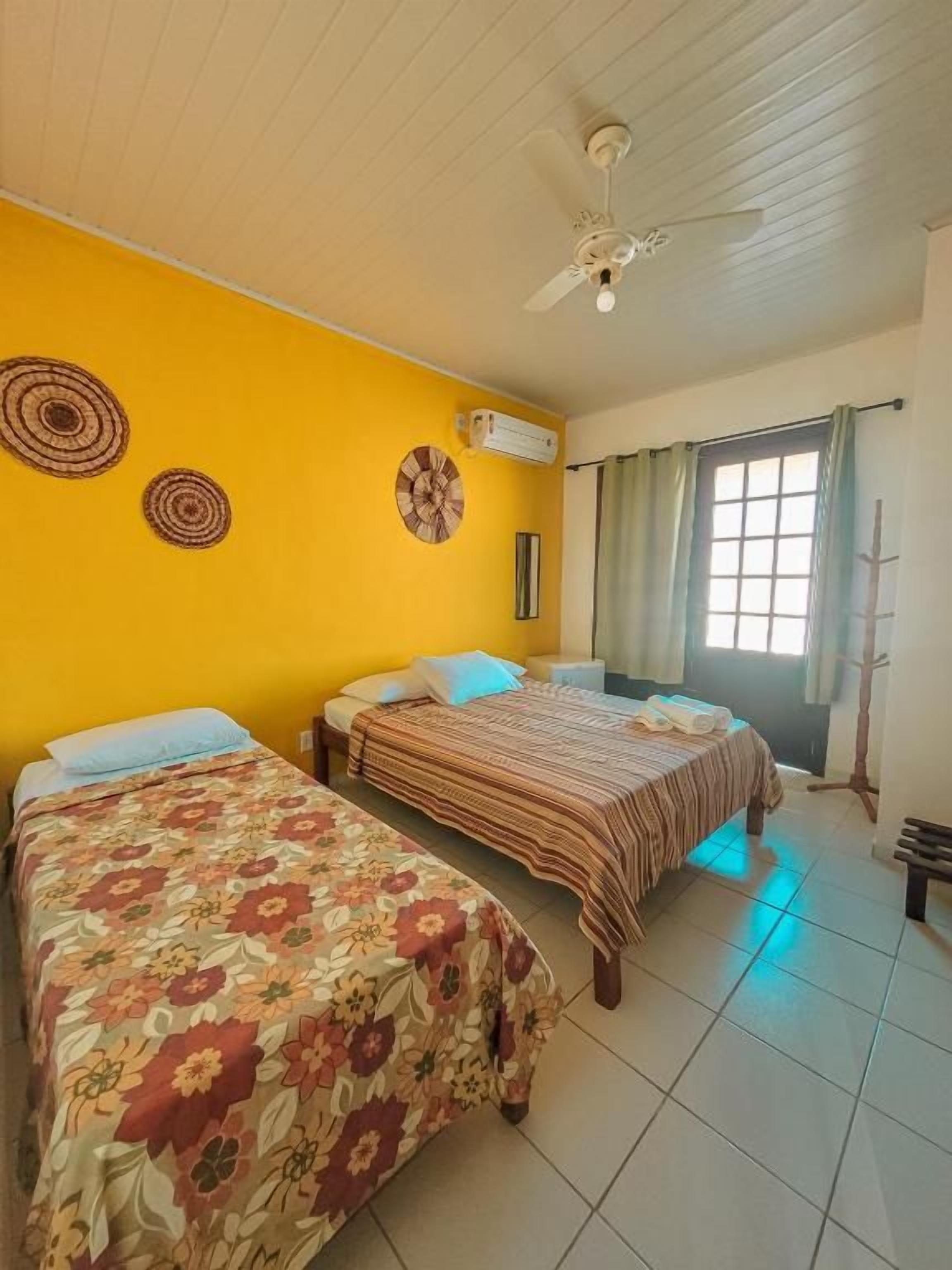 A hotel room with two beds, a ceiling fan, a window with curtains, and a wall-mounted air conditioner.