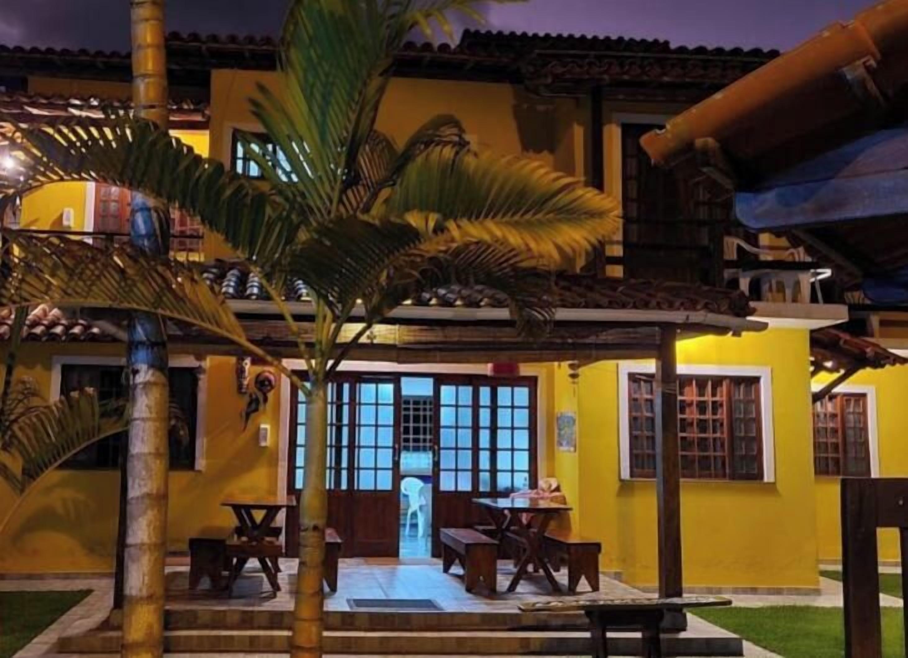 Um edifício amarelo com varanda coberta, porta com painéis de vidro e uma pequena mesa com banco.