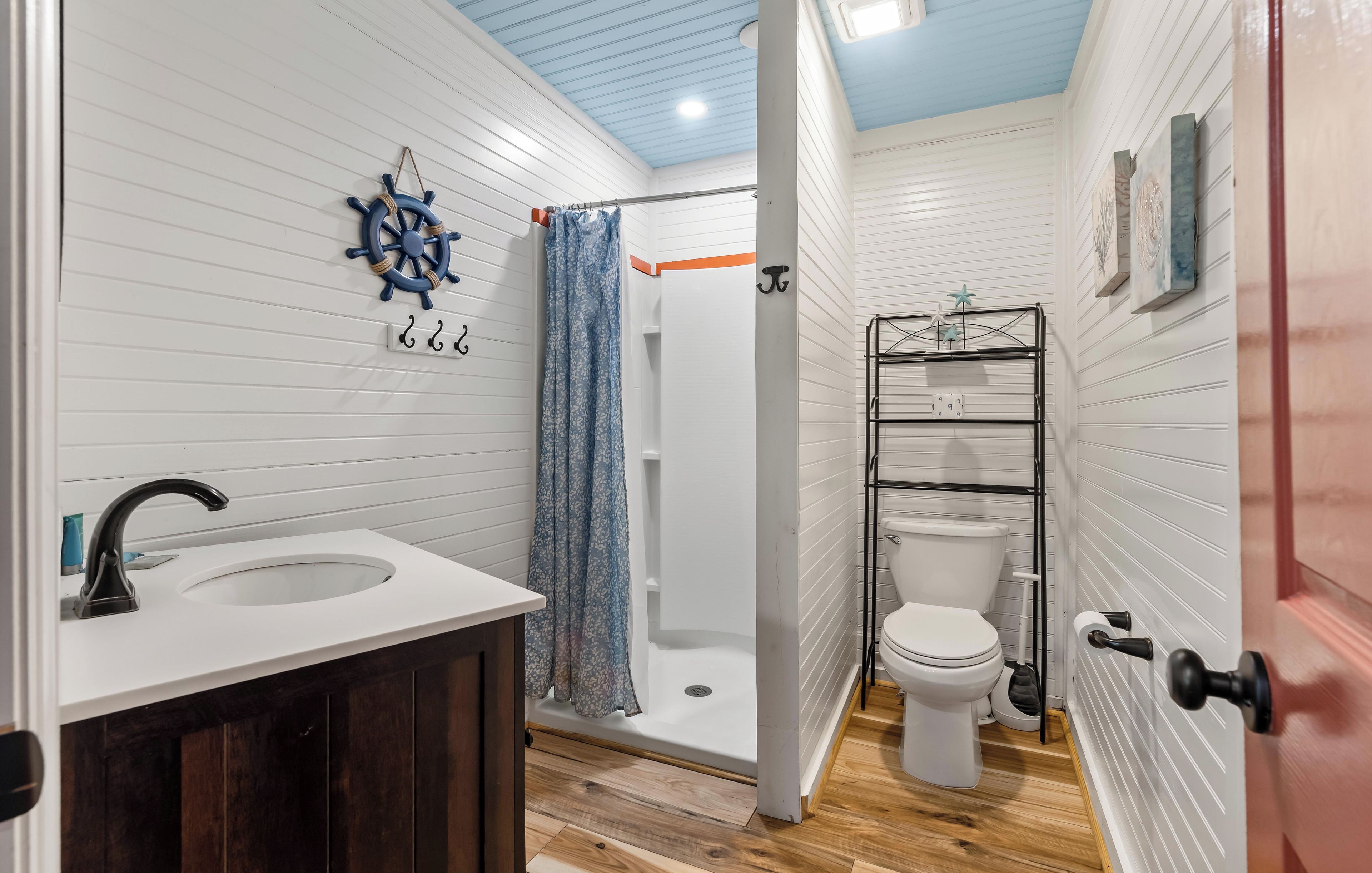 Un baño con lavamanos blanco, inodoro, ducha con cortina y decoración de timón de barco en la pared.