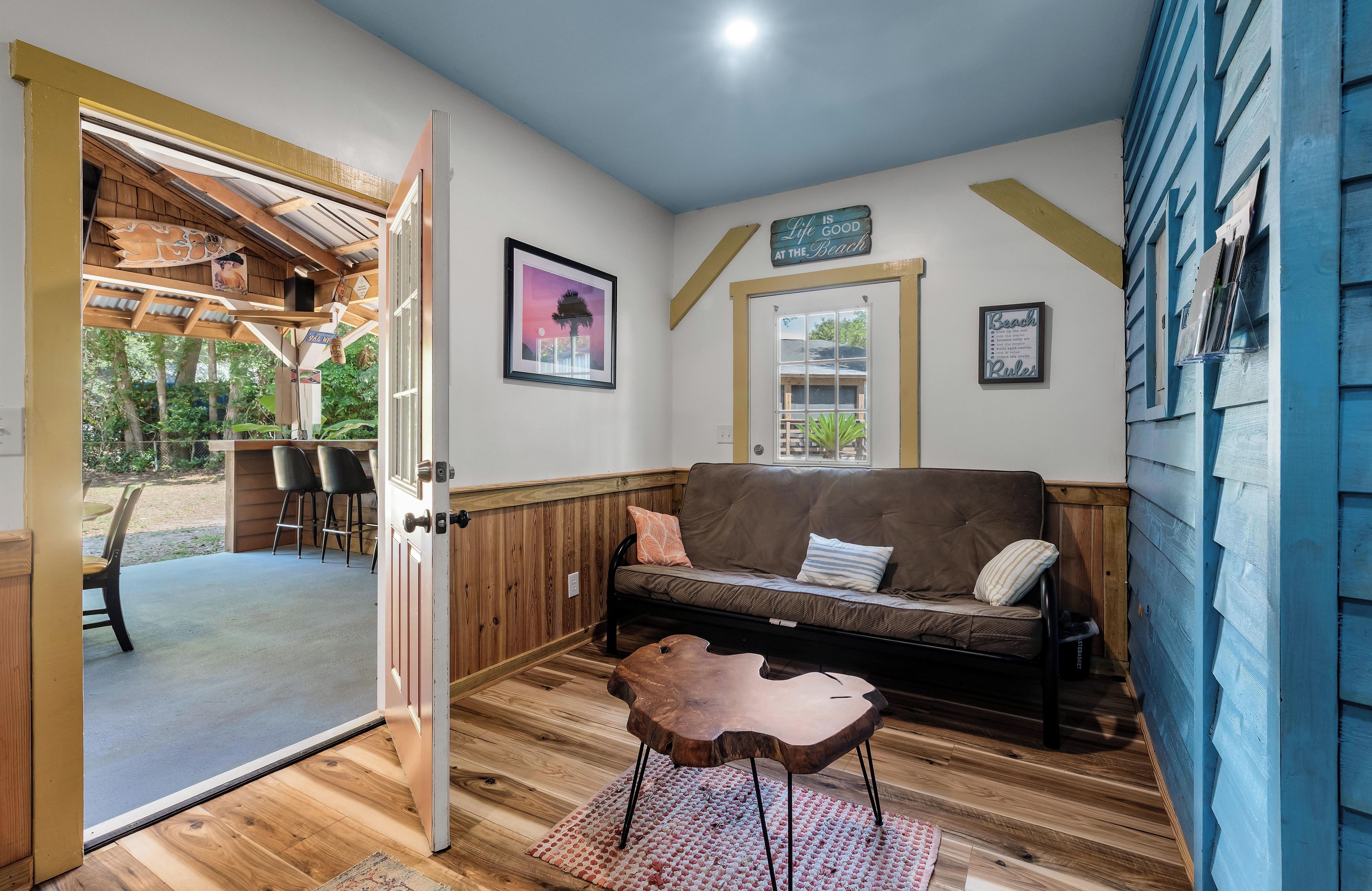Une pièce chaleureuse avec un plancher en bois, un canapé marron, une table basse en bois et une porte donnant sur un espace extérieur aménagé avec des chaises.