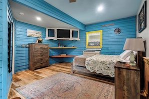 A room with blue walls, a wooden dresser, a bed with a floral bedspread, a small table with a lamp, and a clock on the wall. - Quiet and cozy Coastal Cottage (Charleston)
