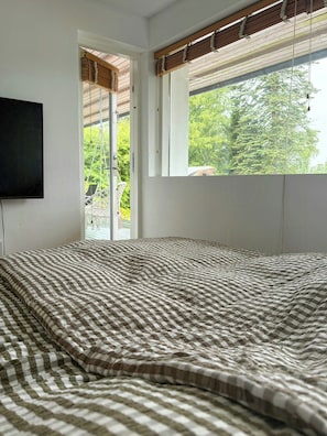 A bedroom with a bed, a television, a window with blinds, and a view of greenery outside. - Private summer house in nature near Ebeltoft (Ebeltoft)