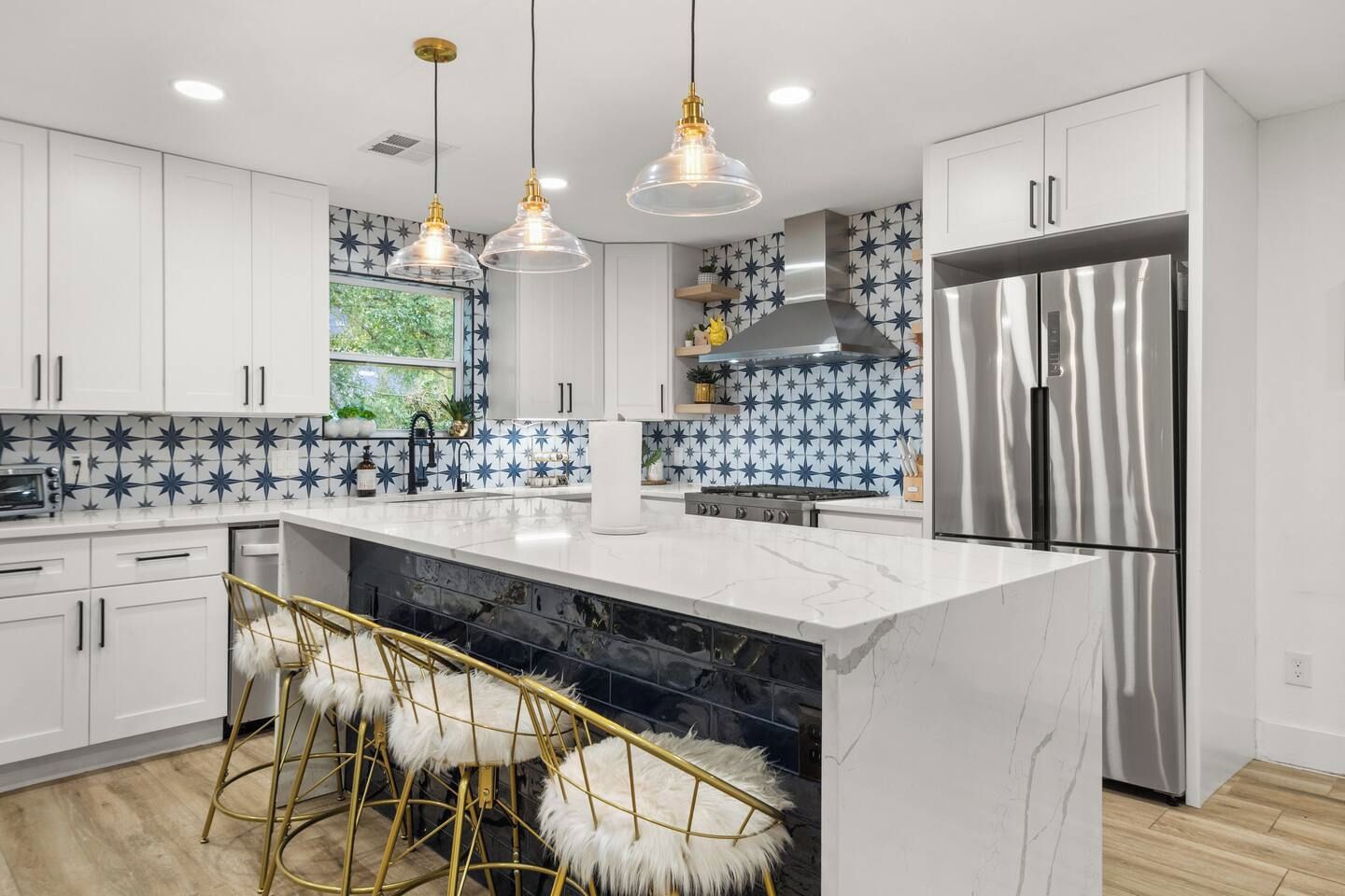 A modern kitchen with white cabinets, a marble island, bar stools, pendant lights, and a stainless steel refrigerator.