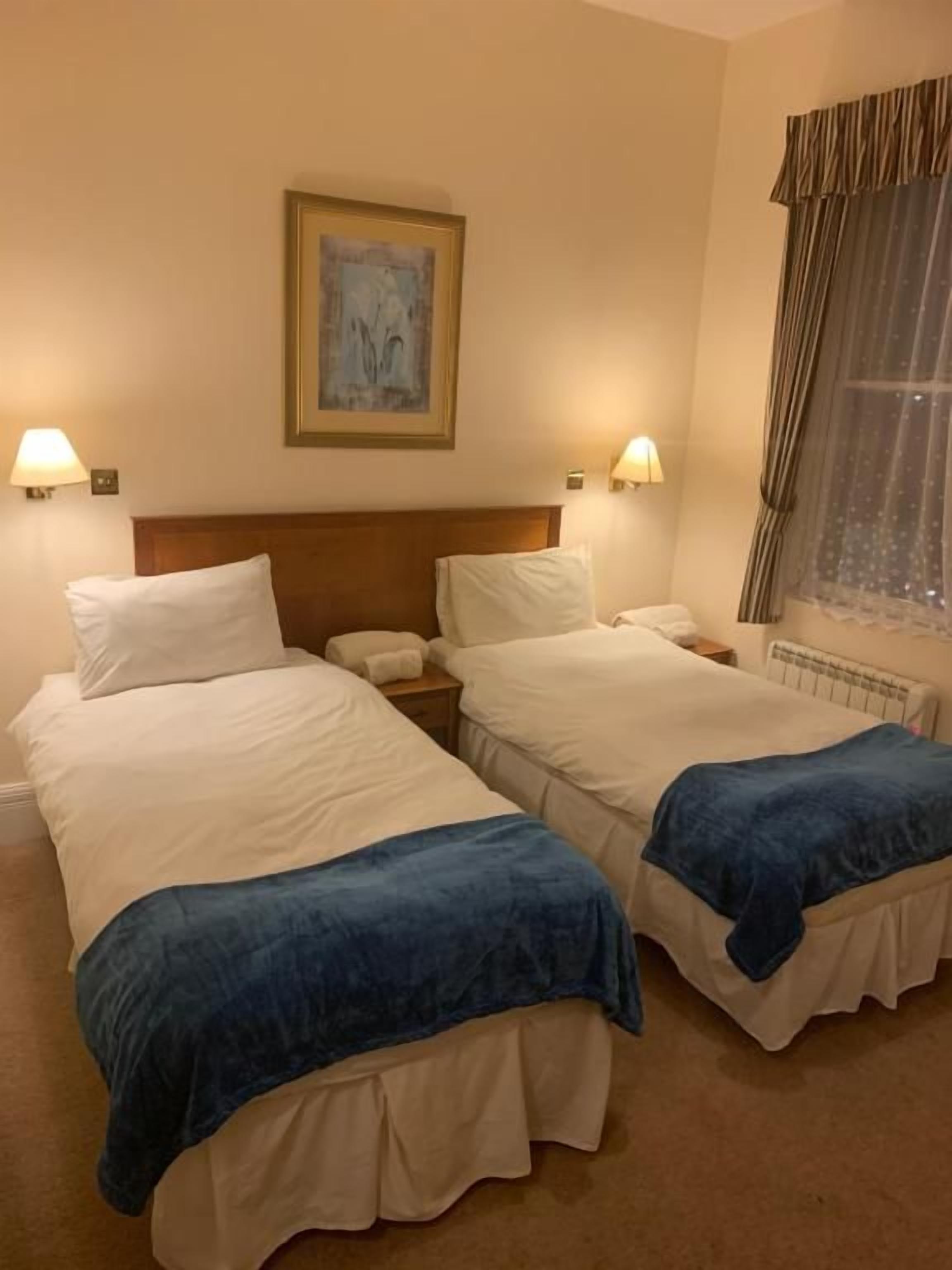 A hotel room with two beds, a headboard, bedside lamps, a painting on the wall, and a window with curtains.